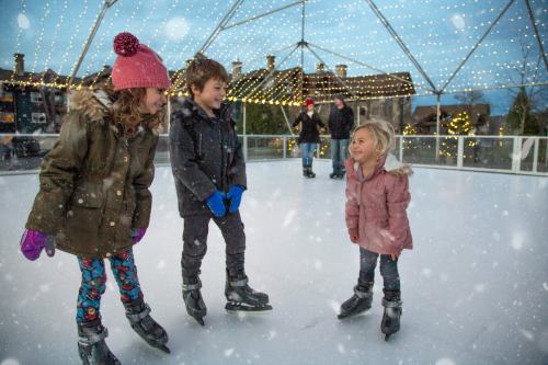 Three children ice skating outdoors, surrounded by festive lights and falling snow, enjoying a winter day.