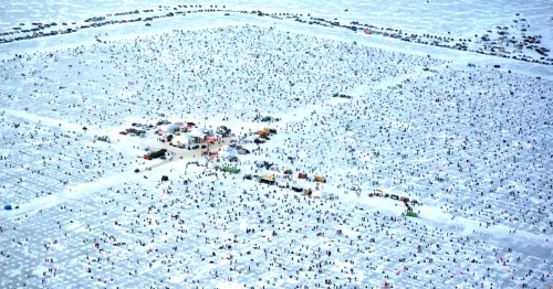 Aerial view of a large crowd at a festival, surrounded by vehicles and tents on a vast, sandy landscape.