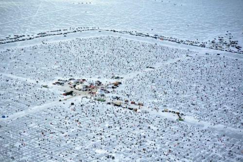 Aerial view of a large crowd on a snowy landscape, with vehicles and tents scattered throughout the area.
