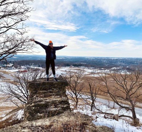 A person stands on a rocky outcrop with arms outstretched, overlooking a scenic landscape of trees and fields.
