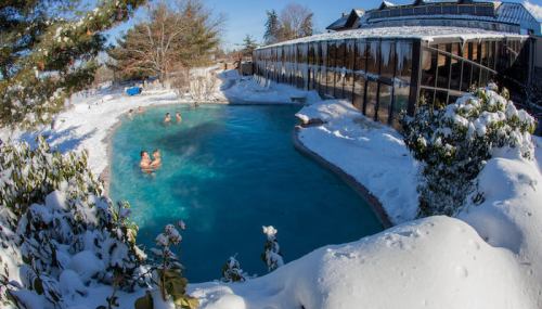 A snowy landscape surrounds a heated pool with people swimming, showcasing a contrast between winter and warm water.