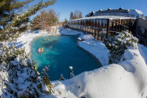 A snowy landscape surrounds a heated pool with people enjoying a swim under a clear blue sky.