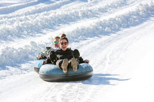 Two people joyfully tubing down a snowy slope, surrounded by white snow and bright sunlight.