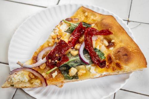 A slice of pizza topped with sun-dried tomatoes, onions, greens, and cheese on a paper plate.