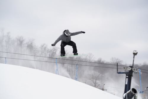 A snowboarder performs a trick on a rail in a snowy landscape, with trees and a ski lift in the background.