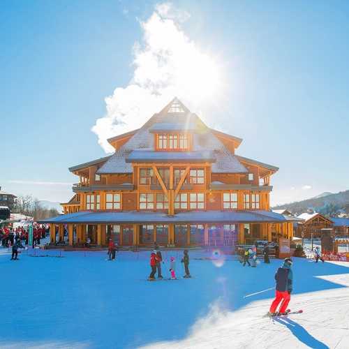 A sunny ski resort with a large wooden lodge and skiers enjoying the slopes in front.