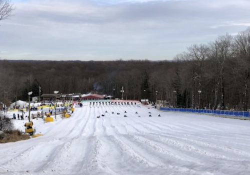 A snowy hill with snow tubing lanes, surrounded by trees and a lodge in the background under a cloudy sky.