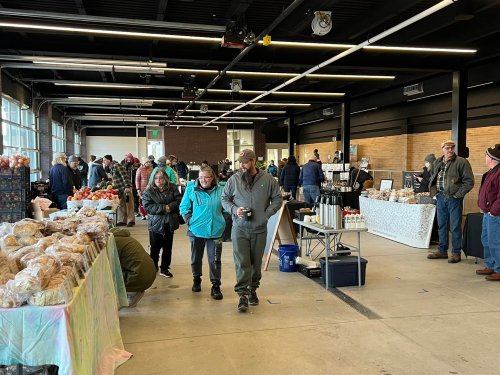 A bustling indoor market with vendors and shoppers browsing various goods, including baked items and crafts.