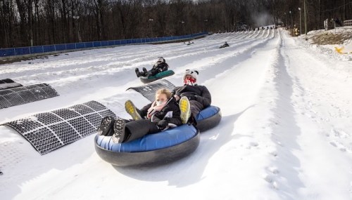 A snowy tubing hill with people sliding down on inflatable tubes, surrounded by trees and a clear blue sky.