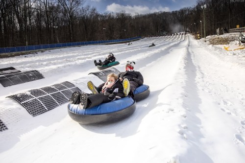 Two people on snow tubes slide down a snowy hill, with others in the background enjoying the winter activity.