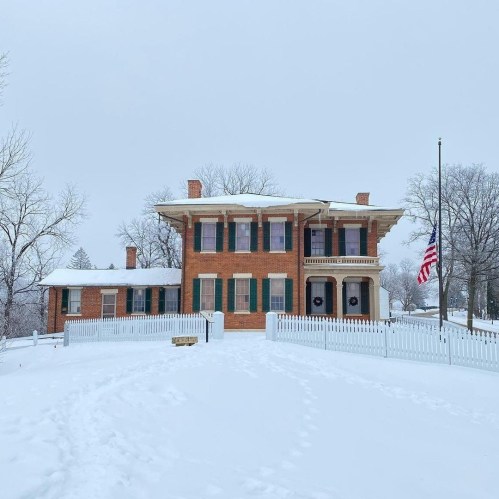 A historic brick house surrounded by snow, with green shutters and a white picket fence, under a gray winter sky.