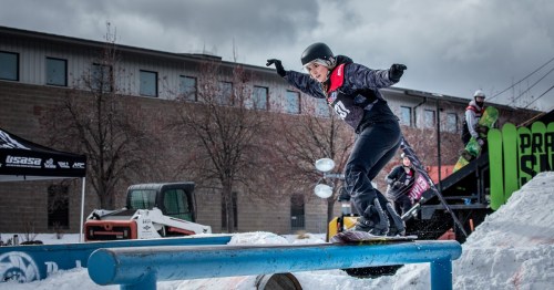 A snowboarder performs a trick on a rail during a competition, with spectators and equipment in the background.