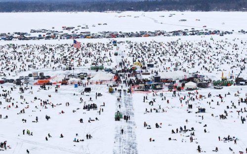 Aerial view of a snowy landscape filled with people ice fishing, surrounded by cars and tents, with an American flag visible.