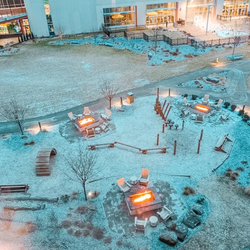 Aerial view of a snowy outdoor area with fire pits, seating, and pathways, surrounded by trees and buildings.