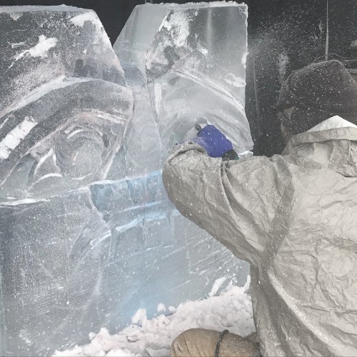 A person chisels an ice sculpture, surrounded by snow and ice blocks, wearing a protective coat and gloves.