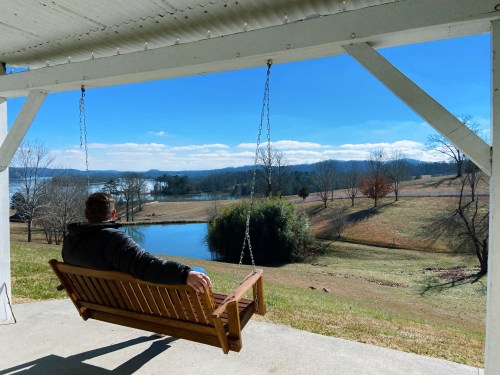 A person sits on a porch swing, overlooking a serene lake and rolling hills under a clear blue sky.