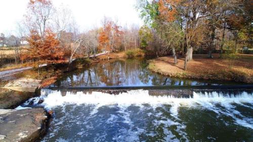 A serene river scene with a small waterfall, surrounded by autumn trees and a calm, reflective water surface.