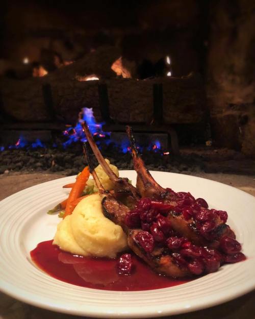 A plate of lamb chops topped with cranberry sauce, served with mashed potatoes and vegetables, with a cozy fireplace in the background.