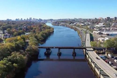 Aerial view of a river with a bridge, surrounded by trees and a city skyline in the background on a clear day.