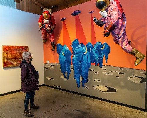 A person observes a colorful mural of blue buffalo and astronauts in a gallery, with UFOs beaming down light.