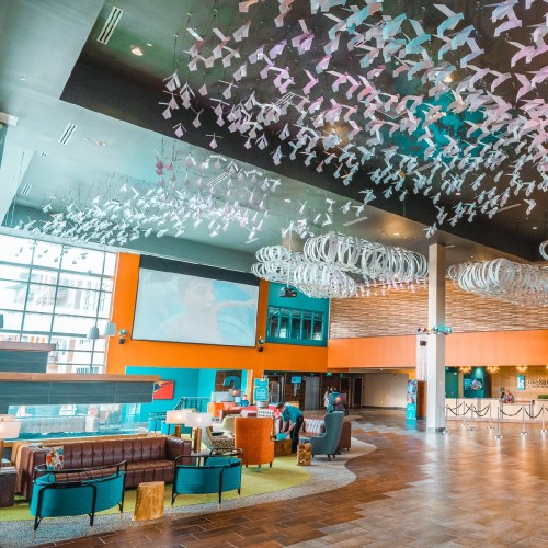 A modern lobby with colorful decor, large windows, and artistic ceiling installations, featuring seating areas and a reception desk.