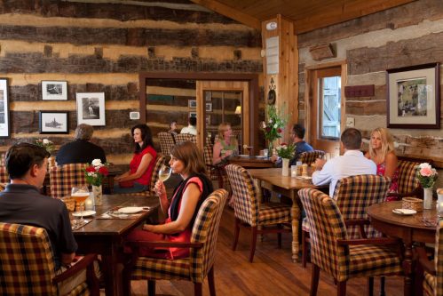 Cozy restaurant interior with wooden walls, patrons dining at tables, and floral centerpieces. Warm, inviting atmosphere.