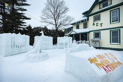 A snowy scene featuring intricate snow sculptures in front of a large house with Greek letters on the facade.