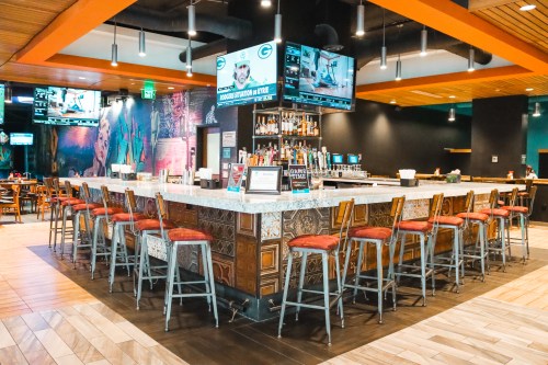 A modern bar with high stools, a marble countertop, and multiple screens displaying sports in a vibrant setting.
