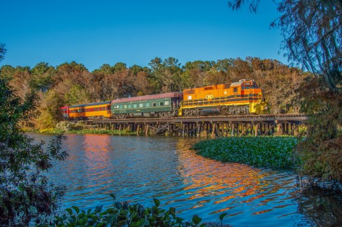 A colorful train crosses a wooden bridge over a calm river, surrounded by lush trees and clear blue skies.