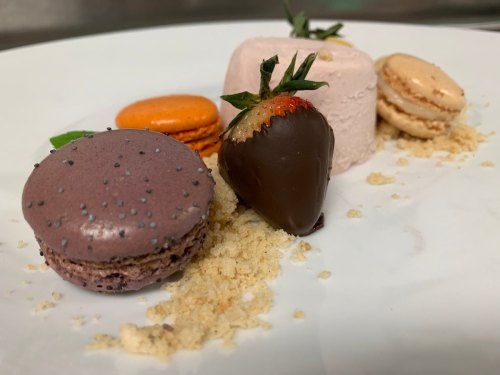 A plate featuring colorful macarons, a chocolate-covered strawberry, and a creamy dessert, garnished with crumbs.