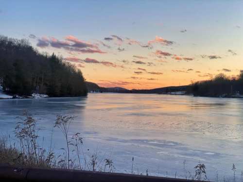 A serene river at sunset, with icy water and trees lining the banks under a colorful sky.
