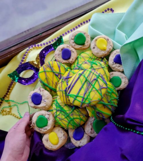 A hand holds a plate of colorful cookies decorated with yellow, green, and purple icing, set on festive fabric.