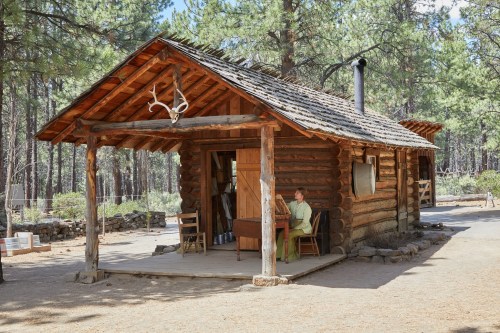 A rustic log cabin surrounded by trees, with a person sitting at a table on the porch.