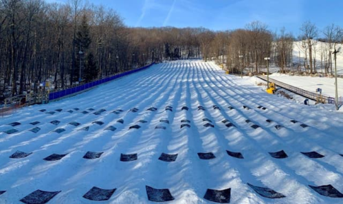 A snowy hill with multiple tubing lanes, surrounded by trees and a clear blue sky.