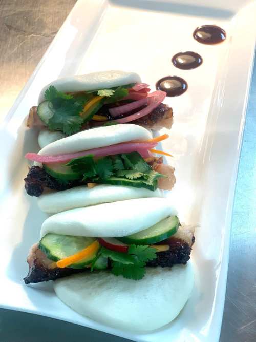 Three bao buns filled with grilled meat, fresh vegetables, and herbs, drizzled with sauce on a white plate.