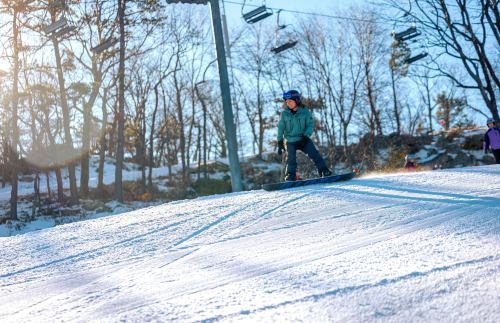 A person snowboarding down a snowy slope with trees and ski lifts in the background, illuminated by sunlight.