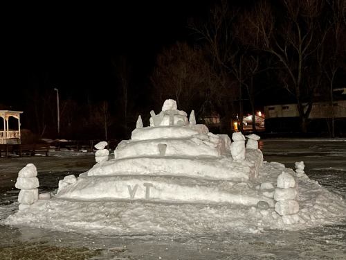 A large snow sculpture resembling a pyramid with smaller snow figures around it, illuminated at night.