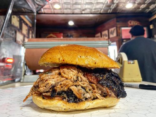 A close-up of a pulled pork sandwich on a bun, with a rustic background featuring a deli counter.