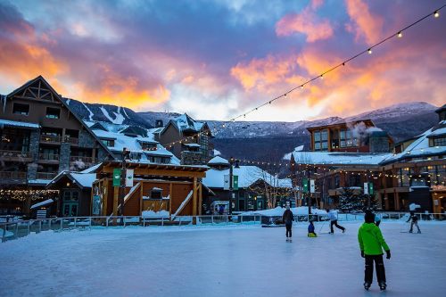 A vibrant sunset over a snowy ice skating rink, surrounded by charming buildings and twinkling lights.