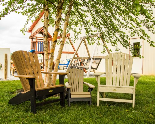 Three Adirondack chairs in varying sizes sit on green grass under a tree, with a playground structure in the background.