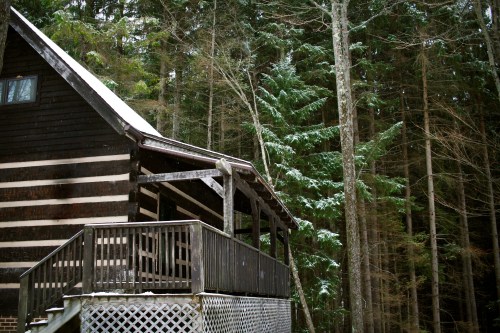 A rustic cabin with a wooden deck, surrounded by tall evergreen trees and a light dusting of snow.