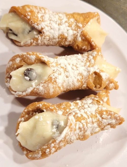 Three cannoli filled with creamy filling and chocolate chips, dusted with powdered sugar on a white plate.