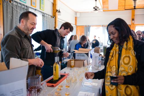 A man pours a drink while a woman samples it at a tasting event, with others mingling in the background.