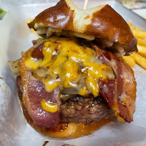 A close-up of a juicy burger with melted cheese, crispy bacon, and a pretzel bun, served with fries on the side.