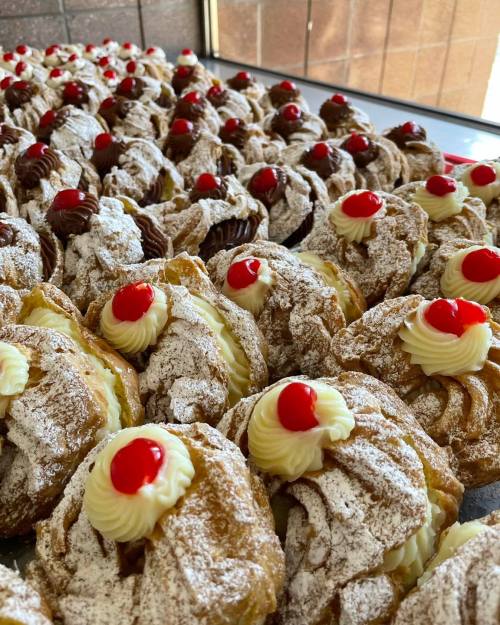A close-up of a tray filled with cream-filled pastries topped with whipped cream and cherries, dusted with powdered sugar.