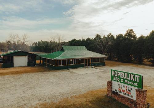 A green-roofed BBQ and market building with a sign, surrounded by trees and a gravel lot.
