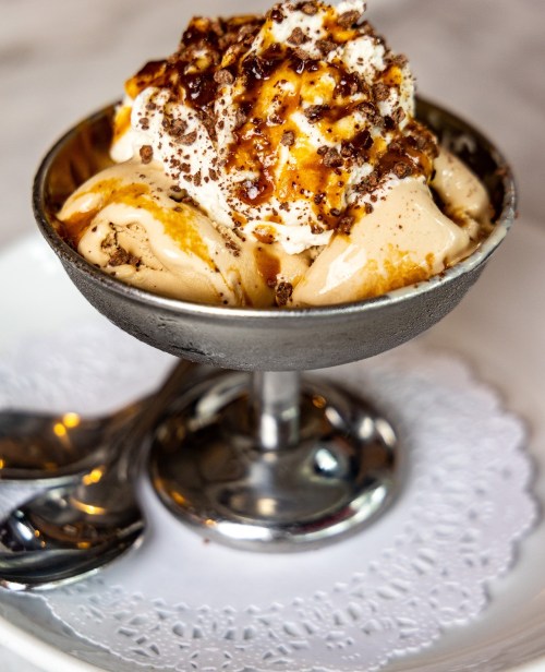 A silver bowl filled with ice cream topped with whipped cream, caramel, and crushed nuts, served on a lace doily.
