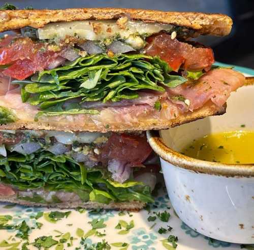 A stacked sandwich with layers of greens, tomatoes, and meats, served with a small bowl of olive oil for dipping.