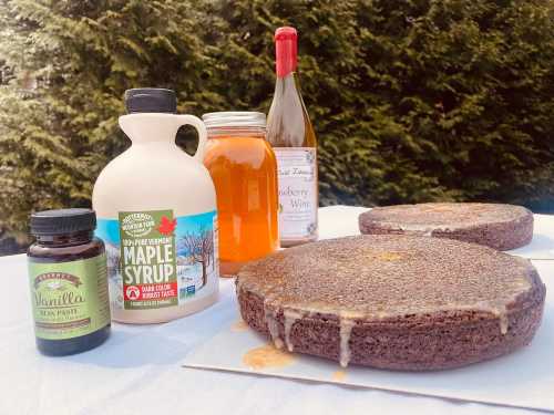 Two round cake layers on a table with maple syrup, vanilla paste, honey, and strawberry wine in the background.