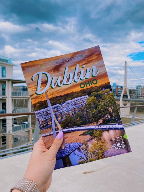 A hand holds a travel guide titled "Dublin, Ohio" against a scenic backdrop of buildings and a cloudy sky.